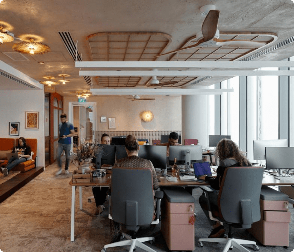 monday.com office: people working at shared desks with monitors beside an orange lounge nook under warm ceiling panels